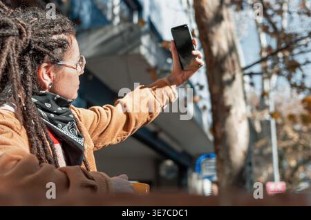 Hispanische Frau mit Dreadlocks, die eine Brille trägt und tagsüber Selfie macht, während sie am Tisch im Café im Freien sitzt Stockfoto