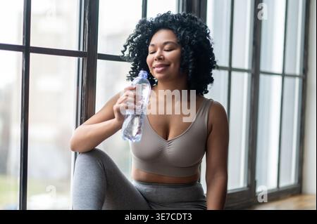 Schwarze Frau mit lockiger Trinkwasserflasche, während sie drinnen auf der Fensterbank sitzt Stockfoto