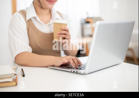 Zugeschnittenes Bild einer asiatischen Frau, die eine Kaffeetasse hält, die mit einem Laptop arbeitet, der zu Hause am Tisch sitzt Stockfoto