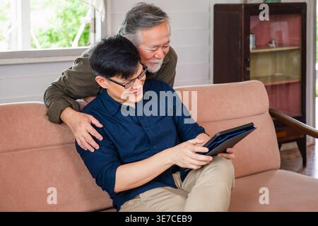 Asiatischer älterer Vater, der sich hinter seinem Sohn mittleren Alters mit einer Brille in der Hand eines Tablets auf der Couch zu Hause liegend Stockfoto