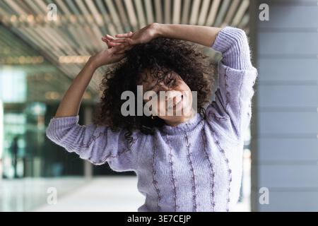 Hispanische Frau mit lockigen Haaren, die tagsüber mit geschlossenen Augen in der Nähe der Glaswand vor dem Gebäude in der Stadt tanzt Stockfoto
