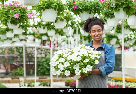 Lächelnde schwarze Frau, die eine Schürze trägt und einen Topf mit Blumen hält, während sie im Gewächshaus steht Stockfoto