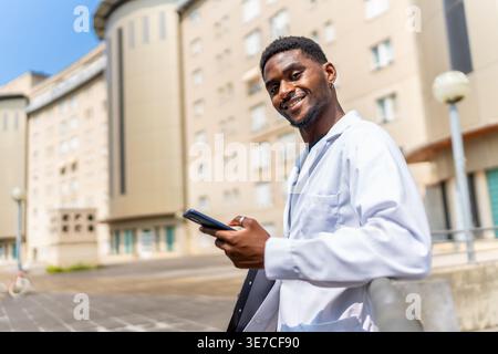 Schwarzer männlicher Arzt, der das Lifestylephone hält und sich an sonnigen Tagen auf den Handlauf vor dem Gebäude lehnt Stockfoto