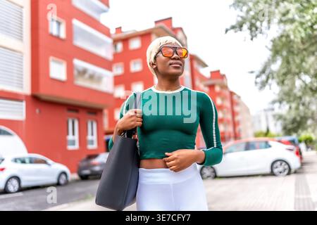 Schwarze Frau mit blonden Haaren, die eine Sonnenbrille trägt und eine Tasche trägt, die tagsüber auf der Straße der Stadt steht Stockfoto