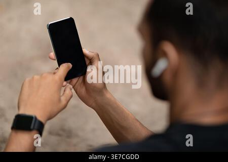 Schwarzer Mann mit Sportbekleidung und Ohrhörern mit Smartphone, während er tagsüber draußen steht Stockfoto
