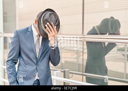 Afroamerikanischer Geschäftsmann, der sein Gesicht hinter seinem Hut versteckt, der in der Stadt in der Nähe einer Glaswand steht Stockfoto