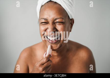 Ältere Frau mit gemischter Rasse, die Badetuch um den Kopf trägt und Spaß bei der Hautpflege hat, während sie vor grauem Hintergrund im Studio steht Stockfoto