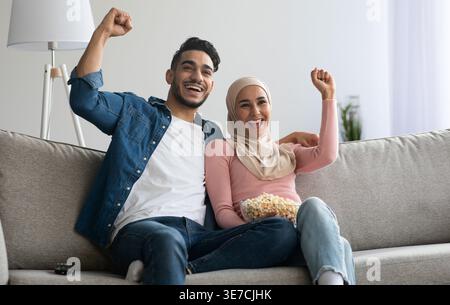 Ein lächelndes Paar aus dem Nahen Osten, das geballte Fäuste beim Popcorn-Essen im Fernsehen hegt, während es zu Hause zusammen auf dem Sofa sitzt Stockfoto