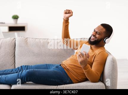 Lächelnder Schwarzer Mann mit geballter Faust, der Musik über kabellose Kopfhörer mit Smartphone hört, während er auf dem Sofa zu Hause im Wohnzimmer liegt Stockfoto
