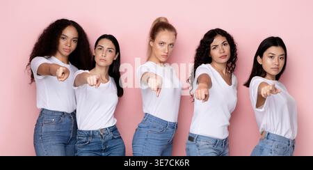 Eine Gruppe von Frauen, die weiße Hemden und Jeans tragen, die mit den Fingern nach vorne zeigen und im Studio vor rosafarbenem Hintergrund stehen Stockfoto