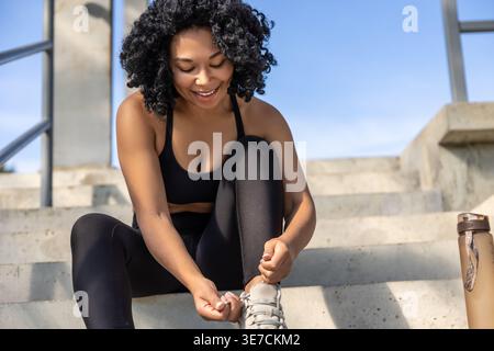 Schwarze Frau mit lockigen Haaren, die an sonnigen Tagen im Stadion auf Treppen sitzt und ein schwarzes Oberteil und Leggings trägt, die ihre Turnschuhe binden Stockfoto