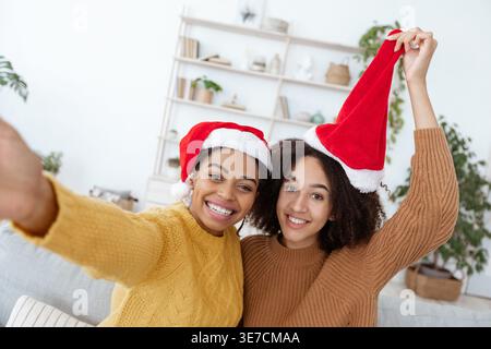 Lächelnd verschiedene weibliche Freunde mit Weihnachtsmützen und Pullover, die zu Hause auf dem Sofa sitzen Stockfoto