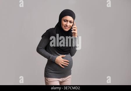 Ängstliche arabische schwangere Frau, die Hijab trägt, ihren Bauch berührt und einen Anruf hat, während sie vor grauem Hintergrund steht Stockfoto