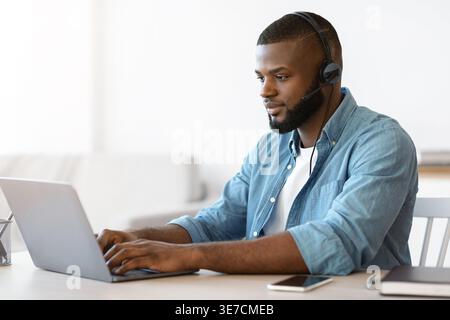 Focus bärtig Schwarzer Freelancer, der ein Headset trägt und tagsüber Videoaufnahmen auf einem Laptop am Tisch im Heimbüro trägt Stockfoto