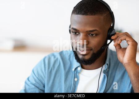 Lächelnder bärtiger Schwarzer Freelancer, der ein Headset trägt, während er im Heimbüro am Tisch sitzt Stockfoto