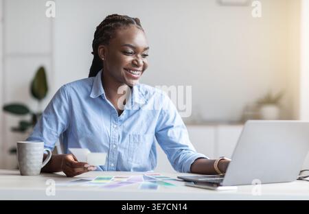 Schwarze Geschäftsfrau mit Flechten, die einen Laptop mit Farbpalette in der Hand hält, während sie im Heimbüro am Tisch sitzt Stockfoto