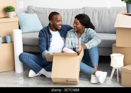 Schwarzes Paar, das Gegenstände in Pappschachtel packt und sich ansieht, während es im Wohnzimmer in der Nähe von ungepackten Kartons zu Hause sitzt Stockfoto