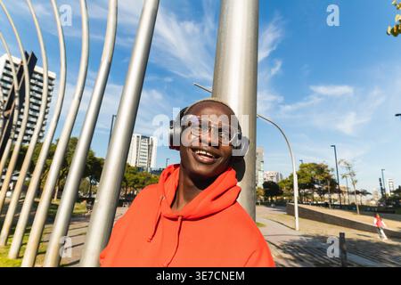 Schwarzer Mann mit kurzen blonden Haaren mit Brille und rotem Kapuzenpullover, der wegblickt, während er am sonnigen Tag im Park auf Metallpfosten steht und sich lehnt Stockfoto
