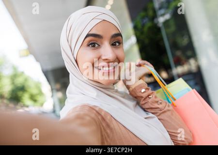 Lächelnde indische muslimische Frau im Hijab, die tagsüber Selfie mit Einkaufstaschen auf der Straße in der Nähe des Einkaufszentrums trägt Stockfoto