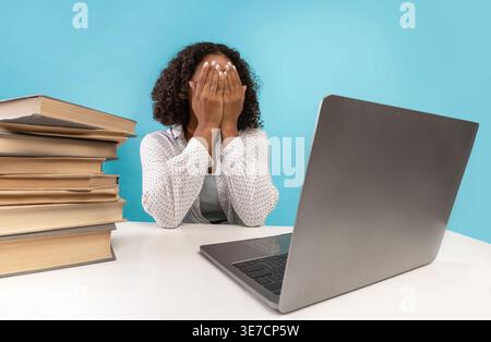 Müde schwarze Teenagerin, die eine Brille mit Laptop trägt, während sie im Studio mit Bücherstapeln vor blauem Hintergrund am Tisch sitzt Stockfoto
