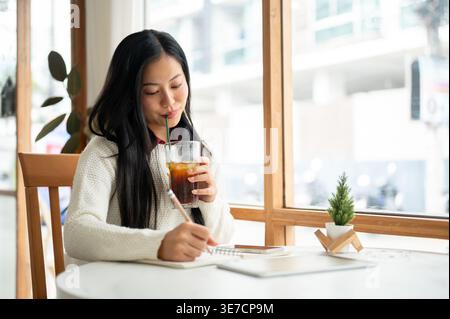 Thailändische Frau mit langen Haaren in Strickjacke, die Notizen macht und Eistee trinkt, während sie mit Smartphone und Tablet im Café am Tisch sitzt Stockfoto