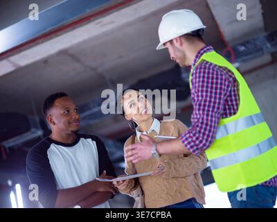 Ein lächelndes Paar aus dem Nahen Osten und ein schwarzes Paar sprechen mit einem männlichen kaukasischen Ingenieur, der ein Tablet hält und über die Planung eines Hauses auf einer Baustelle diskutiert Stockfoto