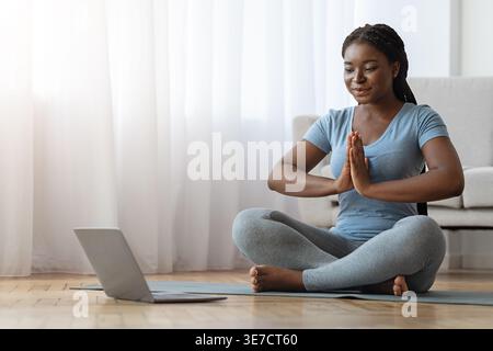 Schwarze Frau mit Online-Meditations-Yoga-Kurs, die in Namaste vor Laptop im Wohnzimmer zu Hause sitzt Stockfoto