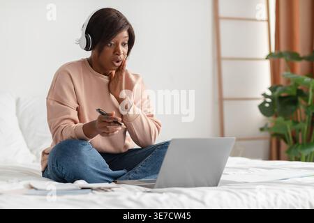 Schockierte Schwarze Frau, die Kopfhörer trägt, das Gesicht berührt, während sie Videoanrufe auf einem Laptop hält, der den Stift hält und mit überkreuzten Beinen auf dem Bett zu Hause im Schlafzimmer sitzt Stockfoto