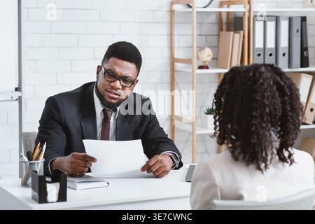Schwarzer männlicher HR-Manager liest den Lebenslauf einer Bewerberin, die während des Vorstellungsgesprächs im Büro am Schreibtisch sitzt Stockfoto