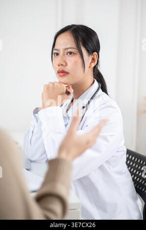 Thailändische Ärztin mit gefesselten Händen am Kinn, die Stethoskop am Hals hält Stift, der weiblichen Patientin zuhört, die ihre Symptome... Stockfoto