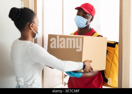 Schwarzer Liefermann mit Gesichtsmaske, der den Paketkasten an weibliche Kunden übergibt, während er während der Pandemie vor der Haustür zu Hause steht Stockfoto