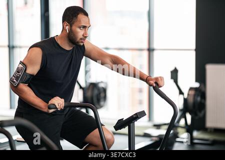 Schwarzer Mann mit Bart, der schnurlose Ohrhörer und Armband trägt und wegschaut, Training mit einem Crosstrainer im Fitnessstudio Stockfoto