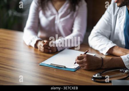 Zugeschnittenes Bild eines schwarzen männlichen Arztes, der ein Rezept für eine Patientin schreibt, die neben ihm in der Klinik sitzt Stockfoto