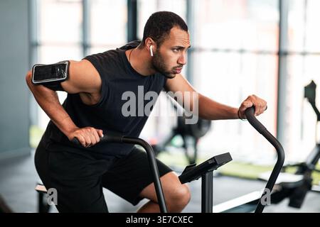 Schwarzer Mann mit Bart, der schnurlose Ohrhörer und Armband trägt und wegschaut, Training mit einem Crosstrainer im Fitnessstudio Stockfoto