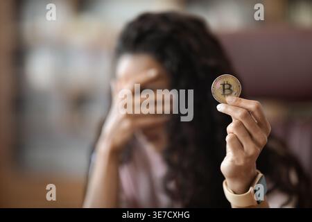 Frau aus dem Nahen Osten mit lockigen Haaren, die Bitcoin zeigt, die ihr Gesicht mit verschwommenem Hintergrund bedeckt, während sie im Büro sitzt Stockfoto