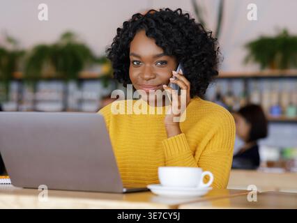 Schwarze Unternehmerin mit lockigen Haaren, die telefoniert am Tisch mit Laptop und Kaffeetasse im Café Stockfoto