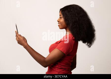 Schwarzer weiblicher Teenager, der ein Smartphone benutzt, während er im Studio vor weißem Hintergrund steht Stockfoto