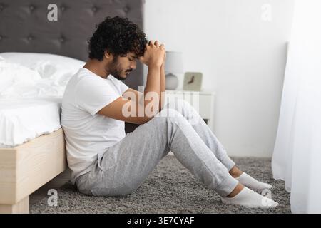 Bärtiger indischer Mann mit lockigem Haar, der die Stirn auf gefalteten Händen lehnt, sitzt auf dem Boden vor dem Bett im Schlafzimmer zu Hause Stockfoto
