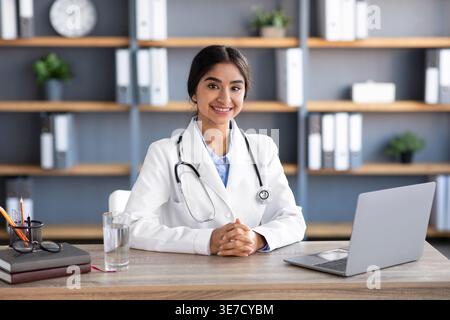 Indische Ärztin mit gefesselten Händen, Stethoskop am Hals am Tisch sitzend mit Laptop und Glas Wasser in der Praxisklinik Stockfoto