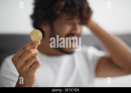 Gestresster bärtiger indischer Mann, der lockiges Haar berührt und goldenes Bitcoin hält, der auf dem Bett zu Hause sitzt Stockfoto
