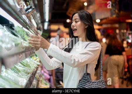 Thailändische Frau mit Schultertasche, die Gemüse aus dem Regal wählt, während sie im Supermarkt steht Stockfoto