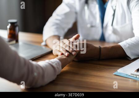Zugeschnittenes Bild eines schwarzen männlichen Arztes mit weißem Labormantel, der die Hand des Patienten während des Sprechens am Tisch hält Stockfoto