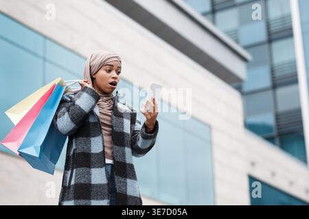 Wow, cooler Rabatt, Verkauf im Online-Shop, Shopping in der Stadt. Überraschte Millennial-Frau islamische afroamerikanerin im Mantel und Hijab mit offenem Mout Stockfoto
