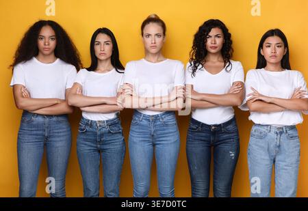 Ernsthafte, vielfältige Gruppe von Frauen in weißen Hemden und Jeans, die mit überkreuzten Armen vor gelbem Hintergrund im Studio stehen Stockfoto