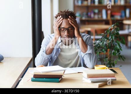 Schwarzer Mann mit Afro und Brille, die den Kopf berührt, schläft am Tisch mit Büchern im Café Stockfoto