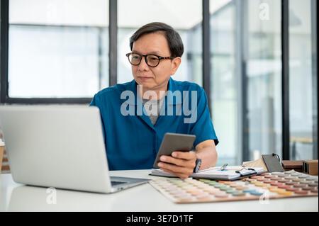 Asiatischer männlicher Innenarchitekt mittleren Alters mit smLifestylephone und Laptop, während er mit Materialien im Büro am Tisch sitzt Stockfoto