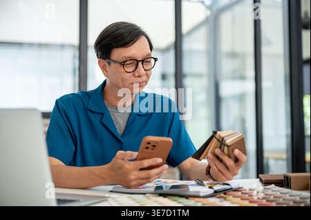 Asiatischer männlicher Innenarchitekt mittleren Alters mit smLifestylephone mit Holzproben am Tisch im Büro Stockfoto