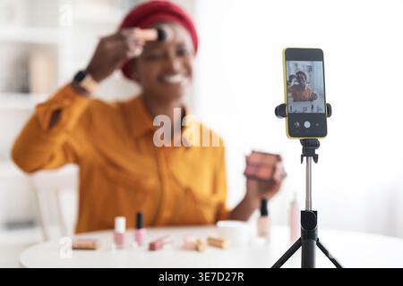 Schwarze Frau mit Kopftuch, die am Tisch sitzt und ein Video aufnimmt, wie sie einen Pinsel hält und zu Hause Make-up aufträgt Stockfoto