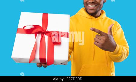 Zugeschnittenes Bild eines lächelnden bärtigen Schwarzen Mannes, der einen Hoodie trägt, der mit dem Finger auf die eingewickelte Geschenkbox zeigt, während er im Studio vor blauem Hintergrund steht Stockfoto