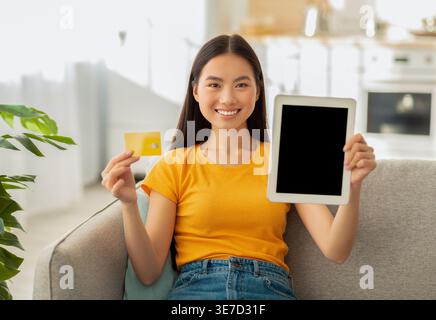 Asiatische Frau zeigt leeren Tablet-Bildschirm und Kreditkarte, die auf Sofa zu Hause im Wohnzimmer sitzt Stockfoto